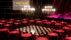 Tables and chairs are arranged all around a black and white checkerboard dance floor under two enormous chandeliers.